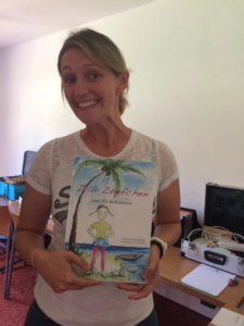 Daniela hält das Buch "Zilli Zöpfchen und die Kokosnuss" lächelnd in die Kamera.