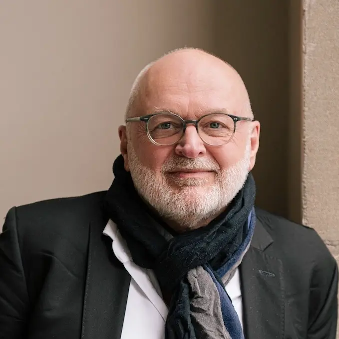 a friendly looking bold man with glasses, a beard, wearing a scarf, a white shirt and a jacket.