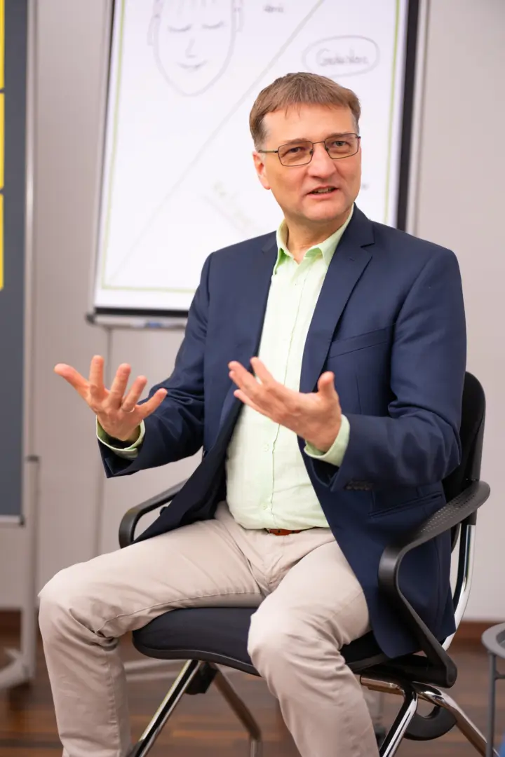Ralph mit Brille und Sacko sitzt und erklärt scheinbar etwas